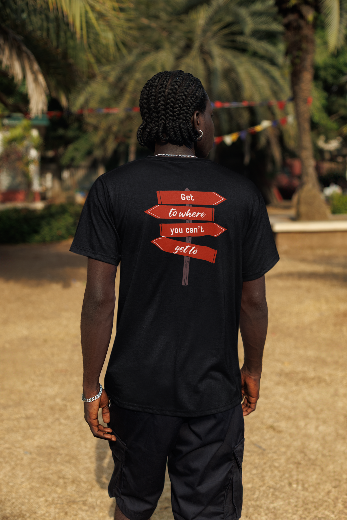A man viewed from behind wearing a black t-shirt with a graphic of three red directional signposts on the back that read 'Go to where you can't get to'. The background is a blurred outdoor scene with palm trees and sandy ground.