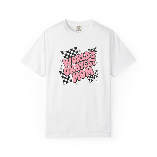 A white graphic t-shirt with the text 'WORLD'S OKAYEST MOM' in pink bubble letters over a black-and-white checkered flag design, paired with light-wash denim shorts in a warm indoor setting.