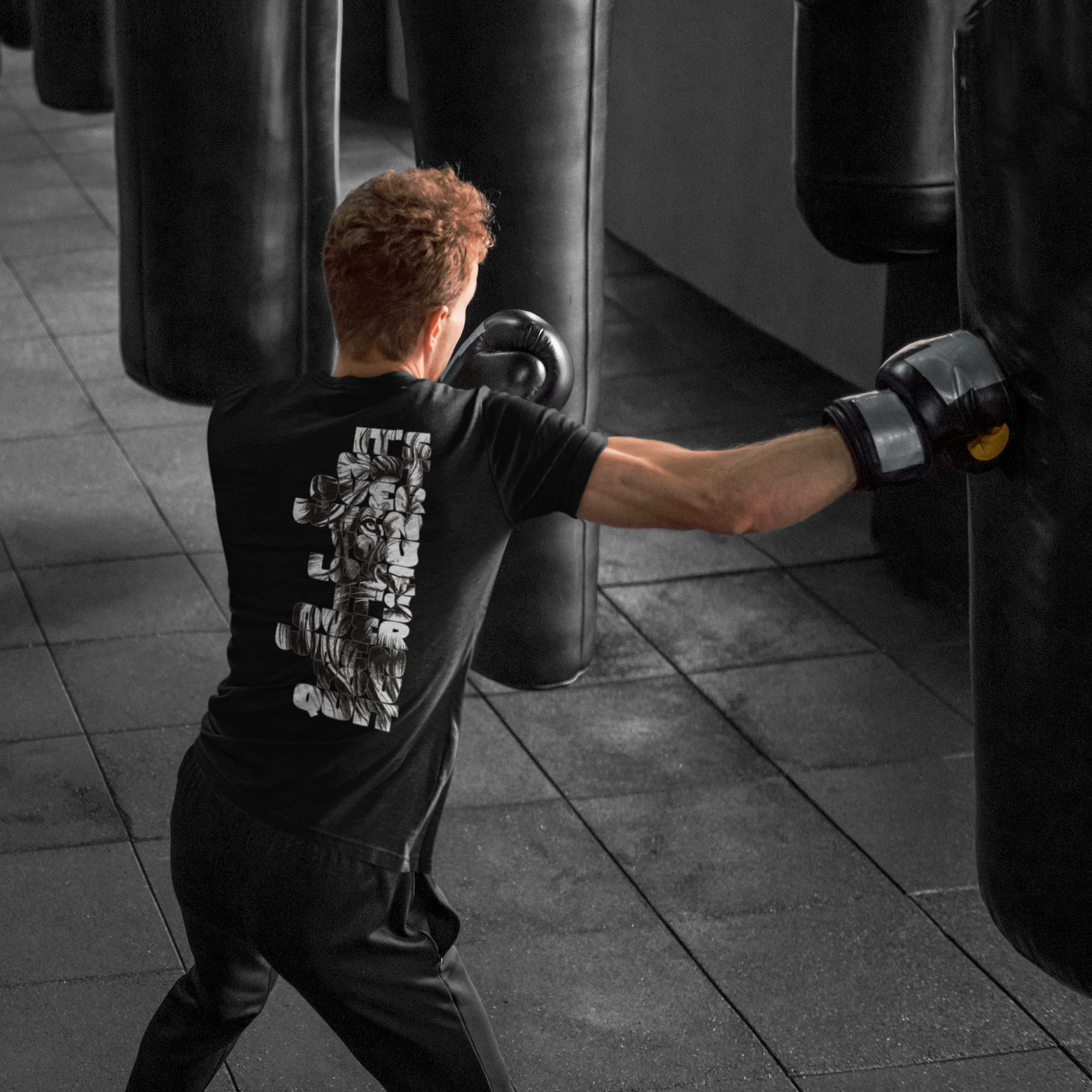 A man in a black t-shirt and black boxing gloves punches a heavy bag in a gym. The back of the t-shirt features a large white graphic print of a boxer.