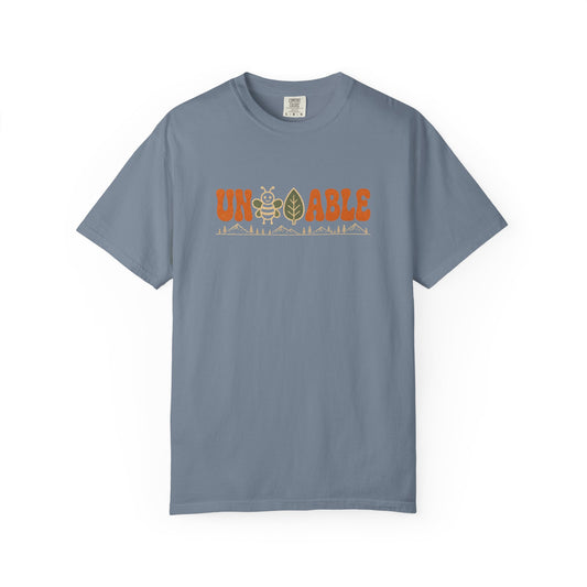 A slate blue crewneck t-shirt with a graphic on the chest reading 'UNBEELEAFABLE' in orange, featuring a bee and a mountain graphic, displayed on a clean white background.