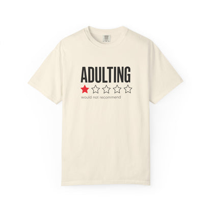 An off-white short-sleeve crewneck t-shirt with a graphic on the chest that reads 'ADULTING' in black bold letters above a one-star red and four-star white outline graphic, with the text 'would not recommend' underneath.