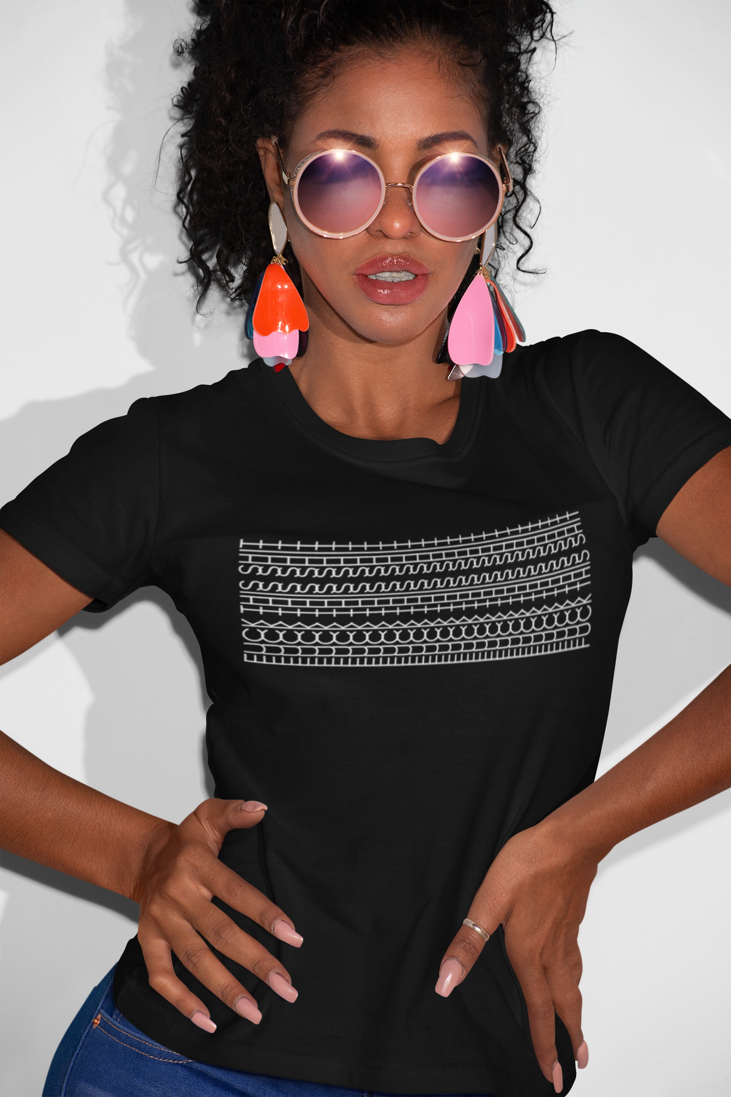 A woman wearing a black short-sleeve t-shirt with a white graphic pattern across the chest, styled with round sunglasses, colorful earrings, and blue jeans against a plain white background.