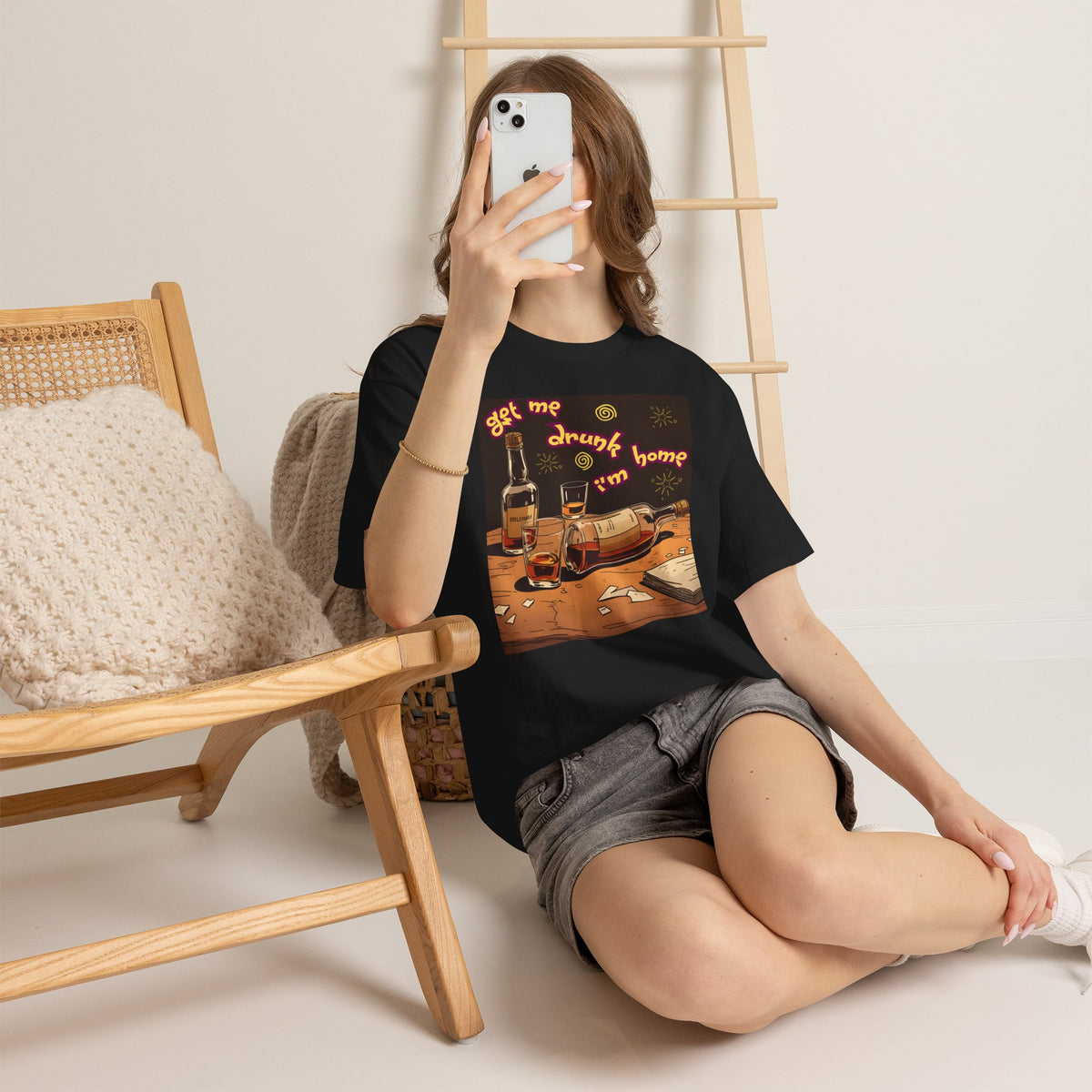 A woman wearing a black short-sleeve crewneck t-shirt featuring a colorful graphic print on the front. The graphic shows an illustration of whiskey bottles, glasses, and shattered glass on a wooden table, with the text 'get me drunk i'm home' in a wavy orange font.