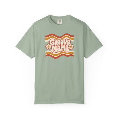 A sage green short-sleeve t-shirt with a retro 1970s-style graphic on the chest reading 'Groovy Mama' in pink bubble letters, set against wavy orange, yellow, and white stripes with small daisy accents.