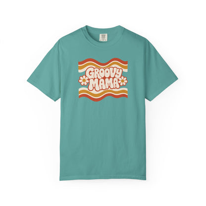 A teal short-sleeve crewneck t-shirt featuring a retro 1970s-style graphic on the chest with wavy orange and yellow stripes, the text 'GROOVY MAMA' in cream bubble letters, and two small orange flowers, displayed on a clean white background.