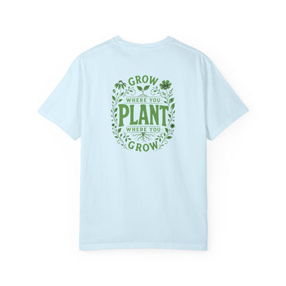 An light blue short-sleeve t-shirt with a green graphic on the back reading 'GROW WHERE YOU PLANT WHERE YOU GROW' surrounded by leaves and clovers, shown against a plain white background.