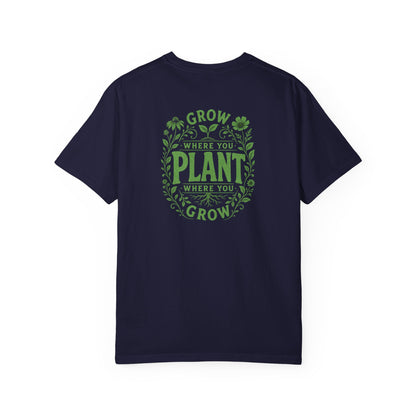A navy blue short-sleeve crewneck t-shirt featuring a green circular graphic on the back with the text 'GROW WHERE YOU PLANT WHERE YOU GROW' surrounded by leaf and clover illustrations.