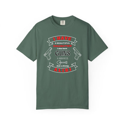 A green short-sleeve crewneck t-shirt with a graphic print on the front. The graphic features the text 'I have a beautiful daughter,i also have guns, a shovel and a strong alibi' in black and red lettering, accompanied by illustrations of two pistols and a shovel.