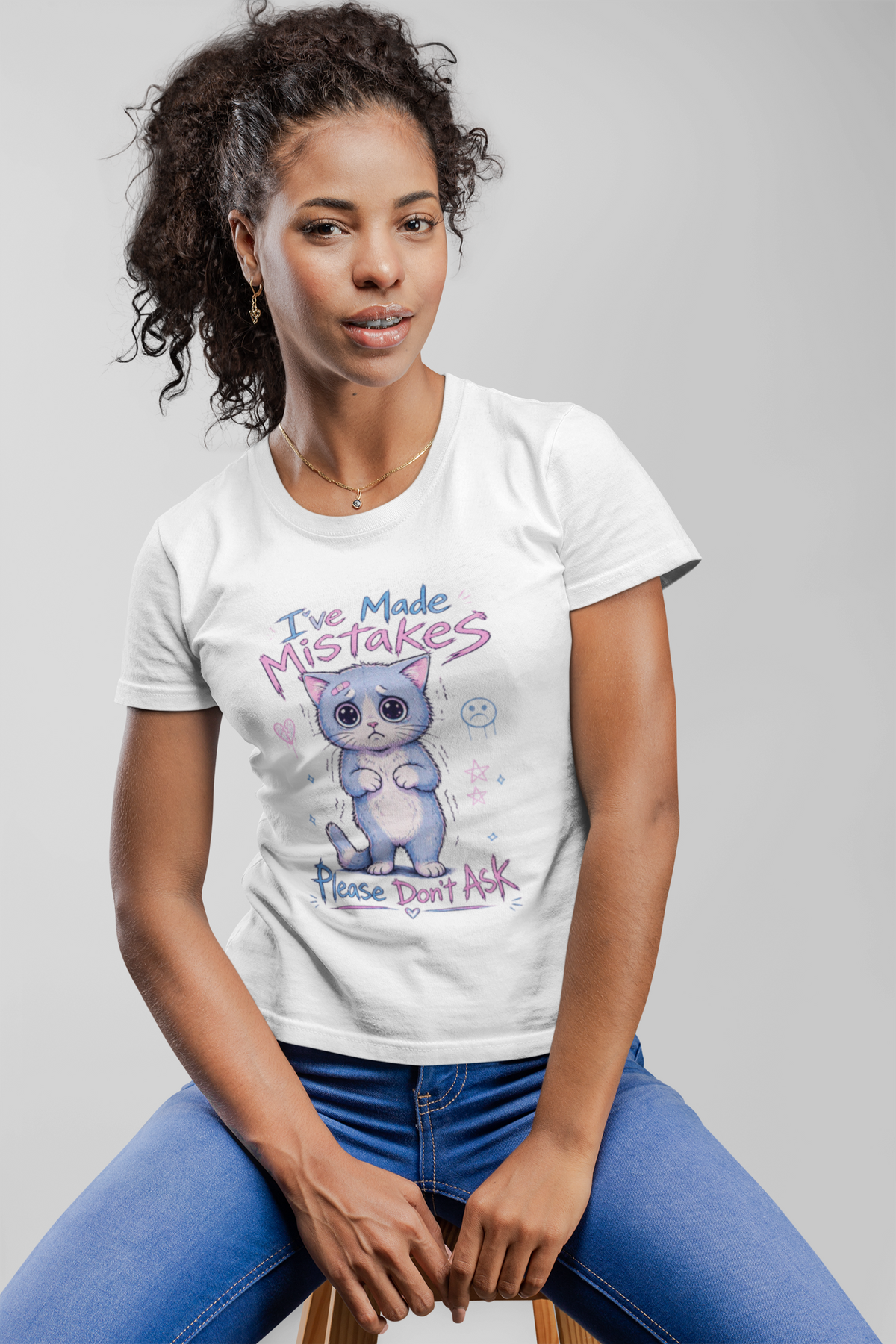 A woman wearing a white graphic t-shirt with a cartoon cat and the text 'I've Made MISTAKES Please don't ask' stands in front of a colorful mural.
