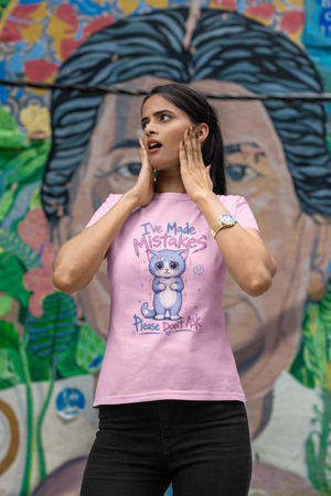 A woman wearing a light pink graphic t-shirt with a cartoon cat and the text 'I've Made MISTAKES Please don't ask' stands in front of a colorful mural.