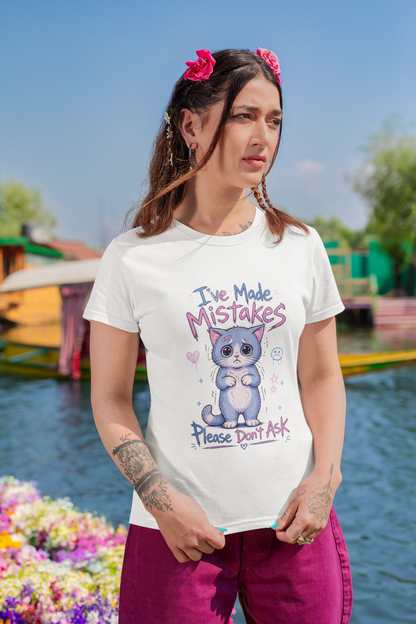 A woman wearing a white graphic t-shirt with a cartoon cat and the text 'I've Made MISTAKES Please don't ask' stands in front of a colorful mural.