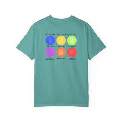 A teal green short-sleeve t-shirt with a colorful graphic on the back. The graphic features six colored circles (blue, yellow, green, purple, orange, red) numbered 1 through 6, with text reading 'IF YOU CAN'T SEE THE NUMBER, YOU: 1 Are Tired 2 Are sad 3 Need Love 4 Need a new bike 5 Are hungry 6 Need a vacation'