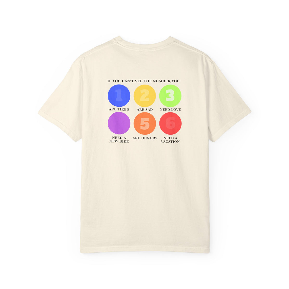 An ivory short-sleeve t-shirt with a colorful graphic on the back. The graphic features six colored circles (blue, yellow, green, purple, orange, red) numbered 1 through 6, with text reading 'IF YOU CAN'T SEE THE NUMBER, YOU: 1 Are Tired 2 Are sad 3 Need Love 4 Need a new bike 5 Are hungry 6 Need a vacation'