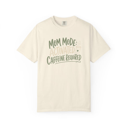 An off-white short-sleeve crewneck t-shirt with a graphic text print reading 'MOM MODE: ACTIVATED CAFFEINE REQUIRED' in dark green and light beige lettering, displayed on a clean white background.