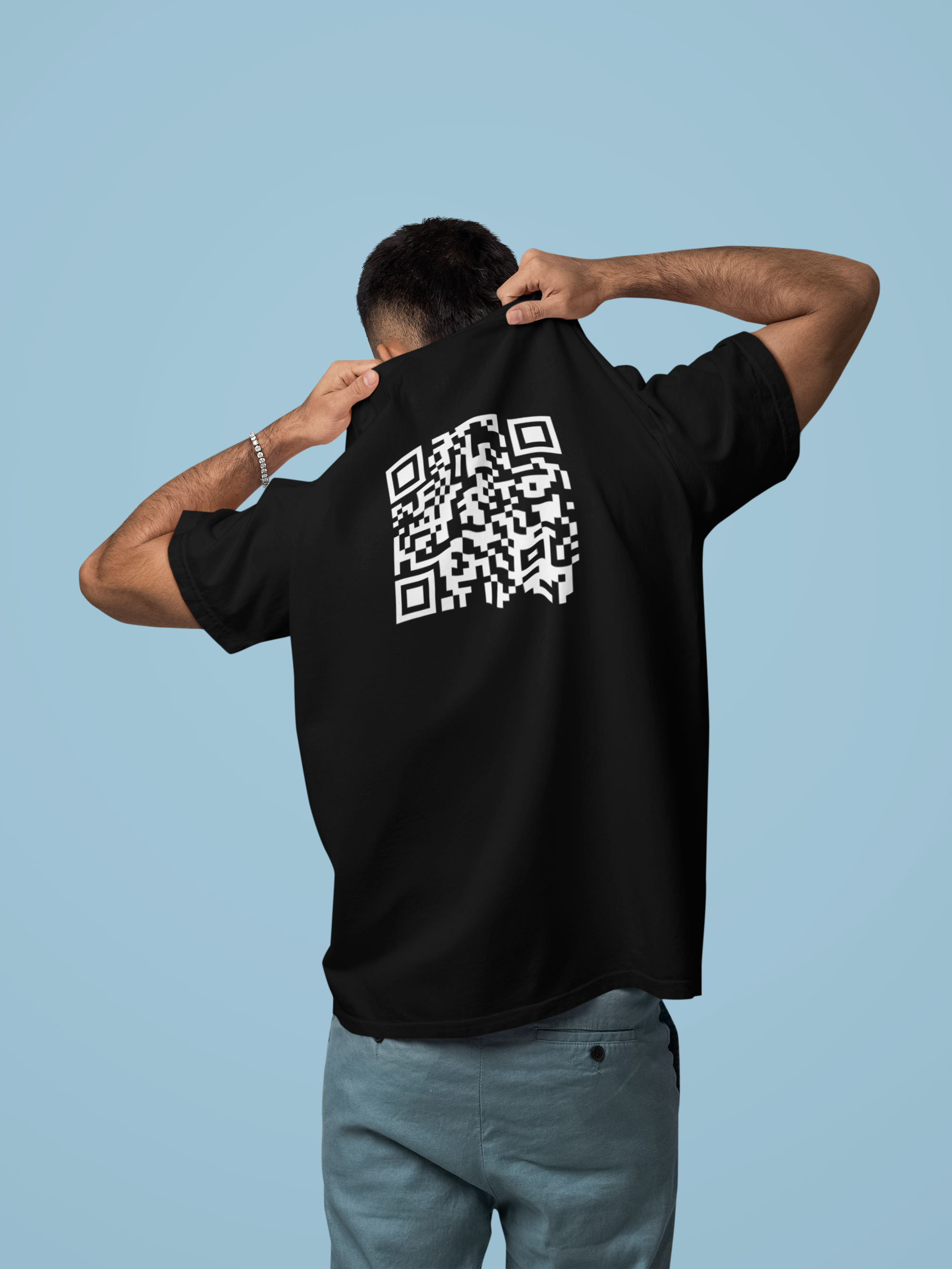 A man viewed from behind wearing a black t-shirt with a large white QR code graphic printed on the back, paired with gray pants, set against a solid light blue background.
