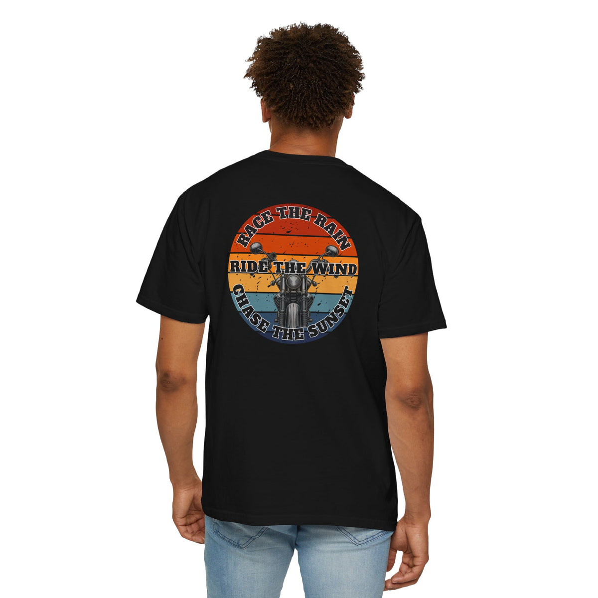 A man wearing a black short-sleeve crewneck t-shirt with a retro circular graphic on the chest. The graphic features a motorcycle silhouette and the text 'RACE THE RAIN', 'RIDE THE WIND', and 'CHASE THE SUNSET' in a distressed sunset color palette of orange, yellow, and blue.