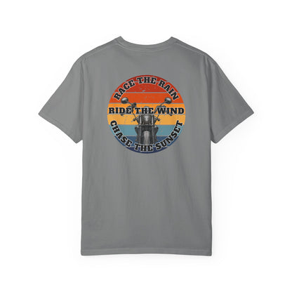 A gray short-sleeve crewneck t-shirt with a retro circular graphic on the chest. The graphic features a motorcycle silhouette and the text 'RACE THE RAIN', 'RIDE THE WIND', and 'CHASE THE SUNSET' in a distressed sunset color palette of orange, yellow, and blue.