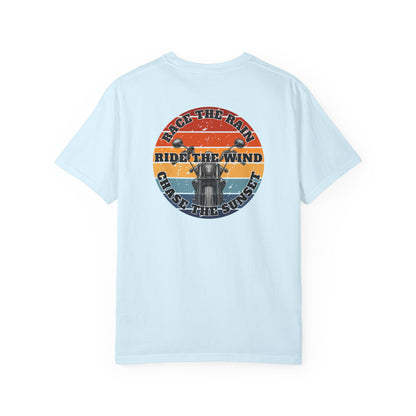 A light blue short-sleeve crewneck t-shirt with a retro circular graphic on the chest. The graphic features a motorcycle silhouette and the text 'RACE THE RAIN', 'RIDE THE WIND', and 'CHASE THE SUNSET' in a distressed sunset color palette of orange, yellow, and blue.