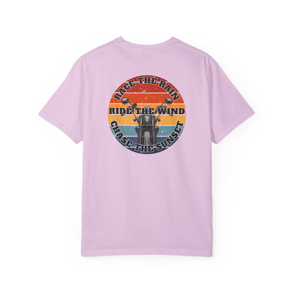 A pink short-sleeve crewneck t-shirt with a retro circular graphic on the chest. The graphic features a motorcycle silhouette and the text 'RACE THE RAIN', 'RIDE THE WIND', and 'CHASE THE SUNSET' in a distressed sunset color palette of orange, yellow, and blue.