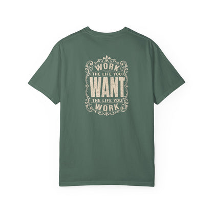 A green short-sleeve crewneck t-shirt with a dark white graphic print on the back. The graphic features the motivational quote 'WORK THE LIFE YOU WANT THE LIFE YOU WORK' in a decorative, vintage-style frame.